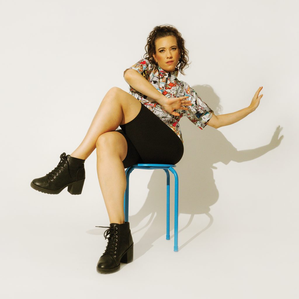 Photograph of artist Caiti Baker sitting down looking at the camera and holding her hands up